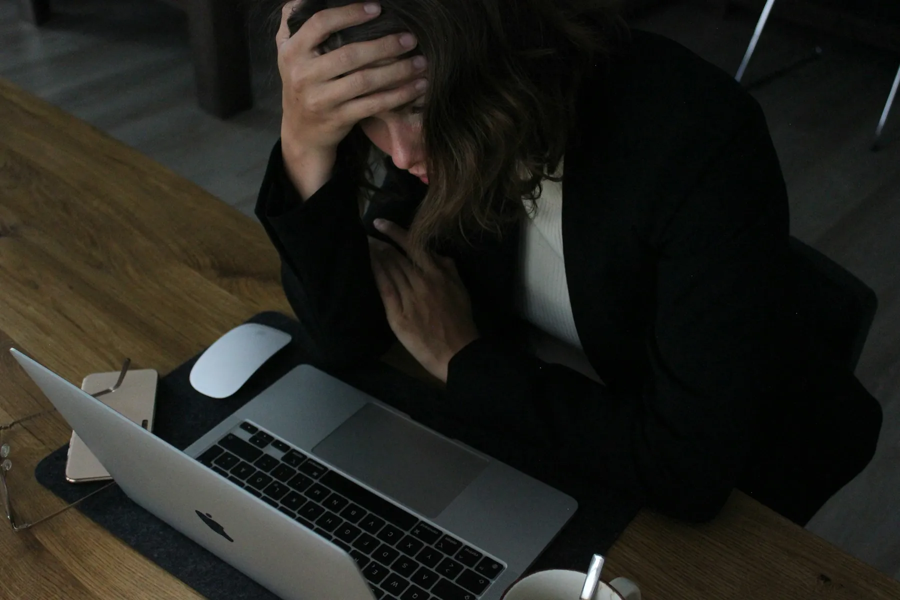 A woman sitting at a laptop in dim light, one hand pressed to her forehead