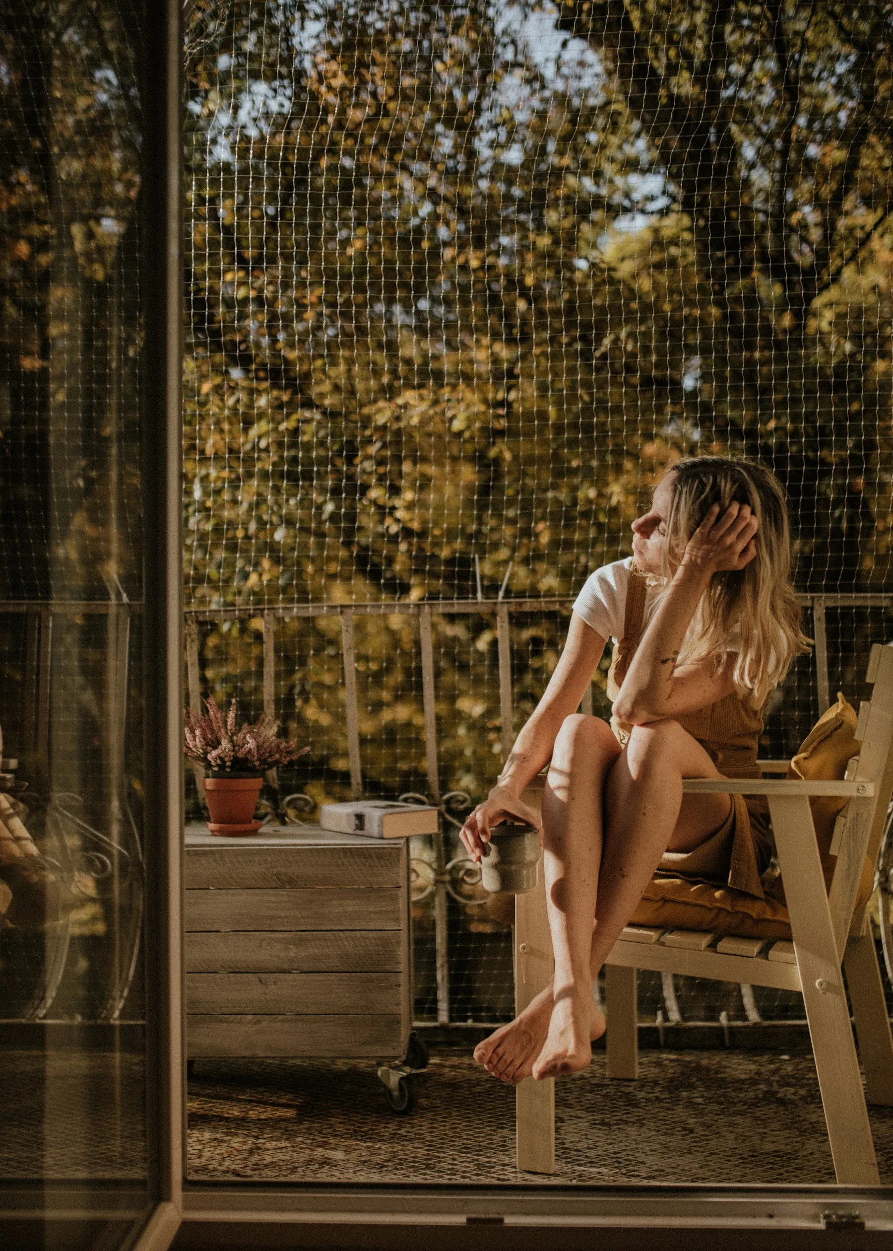A woman sitting barefoot on a balcony chair in autumn light, holding a mug and looking out at the trees