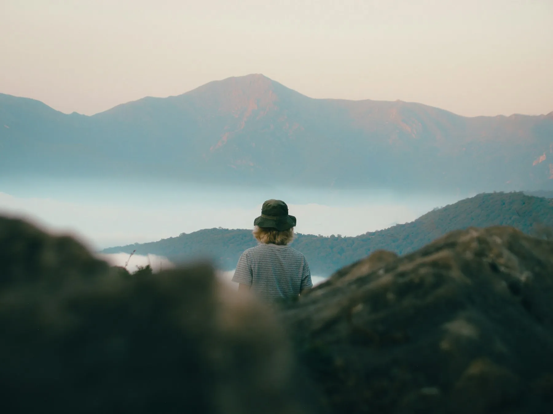 A person in a bucket hat seen from behind, looking out over mist and mountains at first light